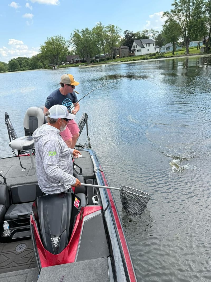 Two anglers on a bass boat reeling in a fish on a calm lake, net ready at the side of the boat