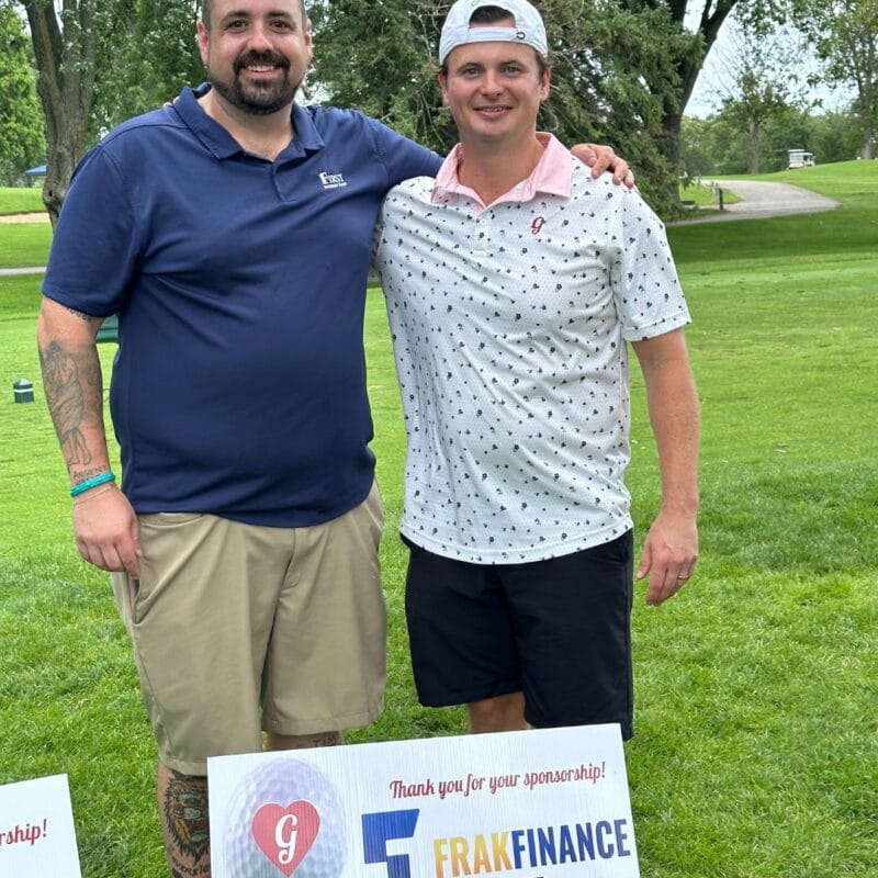 Two men posing on a golf course fairway beside a Frak Finance thank-you sponsorship sign