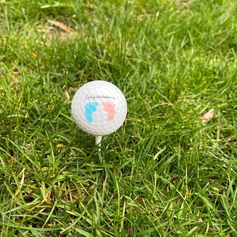 Custom golf ball printed with baby footprints and the word Congratulations, resting on a tee in the grass