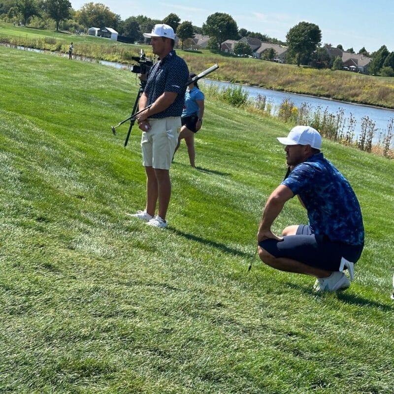 Golfer crouching to read a putt on a hillside fairway while a camera crew films nearby, pond visible in background