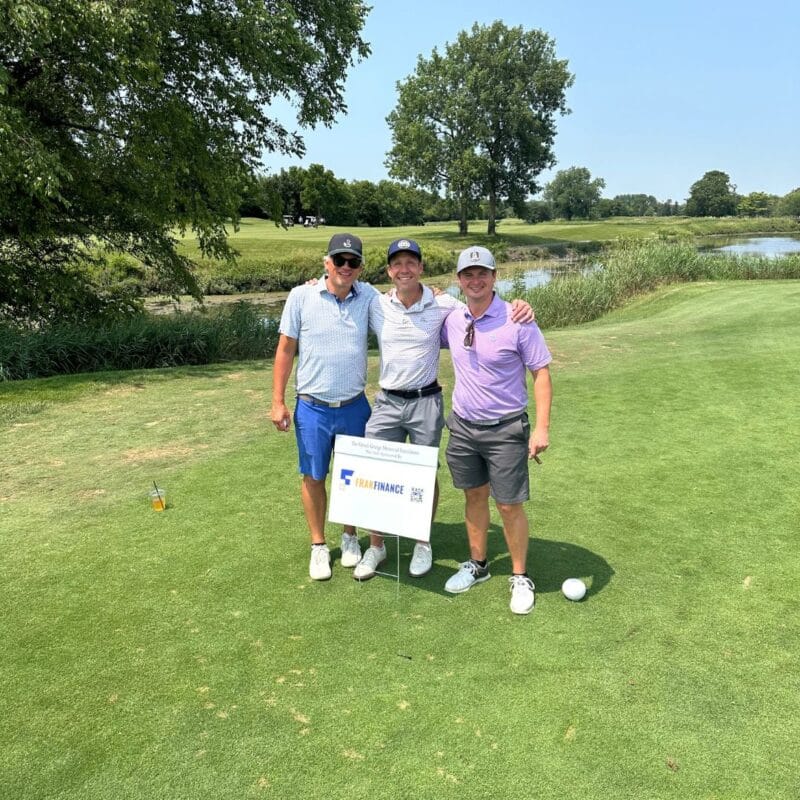 Three golfers posing together on a fairway beside a Frak Finance sponsorship sign at a charity golf tournament