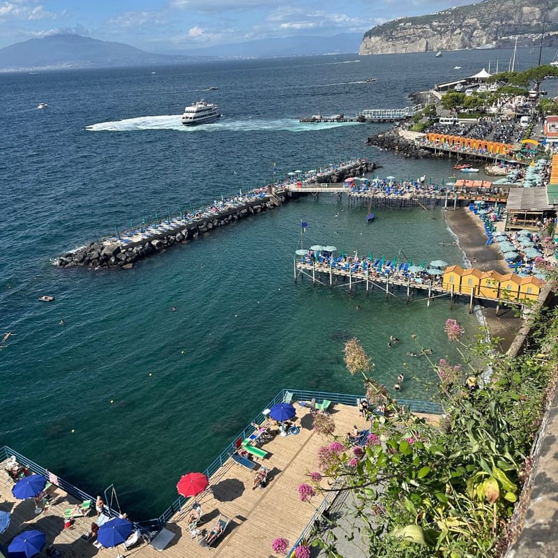 Aerial view of a coastal harbor in Sorrento, Italy, with piers, beach umbrellas, and a ferry crossing the bay
