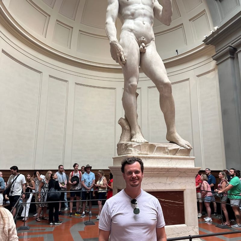 Tom Dillon standing in front of Michelangelo's David at the Galleria dell'Accademia in Florence, surrounded by museum visitors